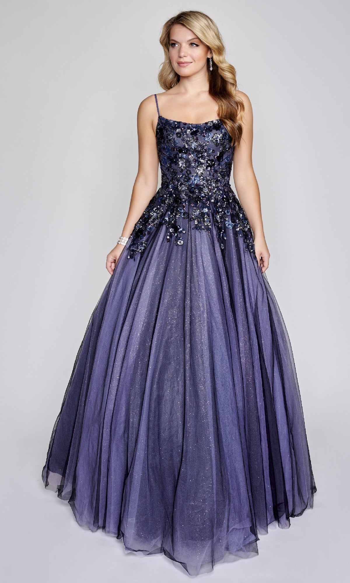 A woman wears the Nina Canacci 3206 prom ball gown—sleeveless, floor-length, purple, with floral bodice embellishments and a glitter tulle skirt—standing against a plain background.
